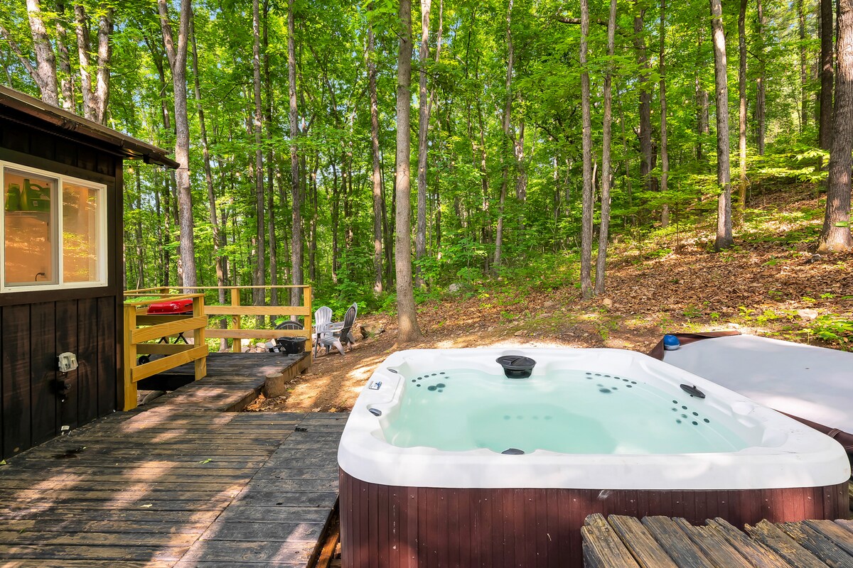 A private hot tub is nestled among lush greenery, surrounded by tall trees. A wooden deck leads to the hot tub, providing a natural and peaceful setting for relaxation. Adjacent seating areas feature Adirondack chairs, inviting serene outdoor moments.