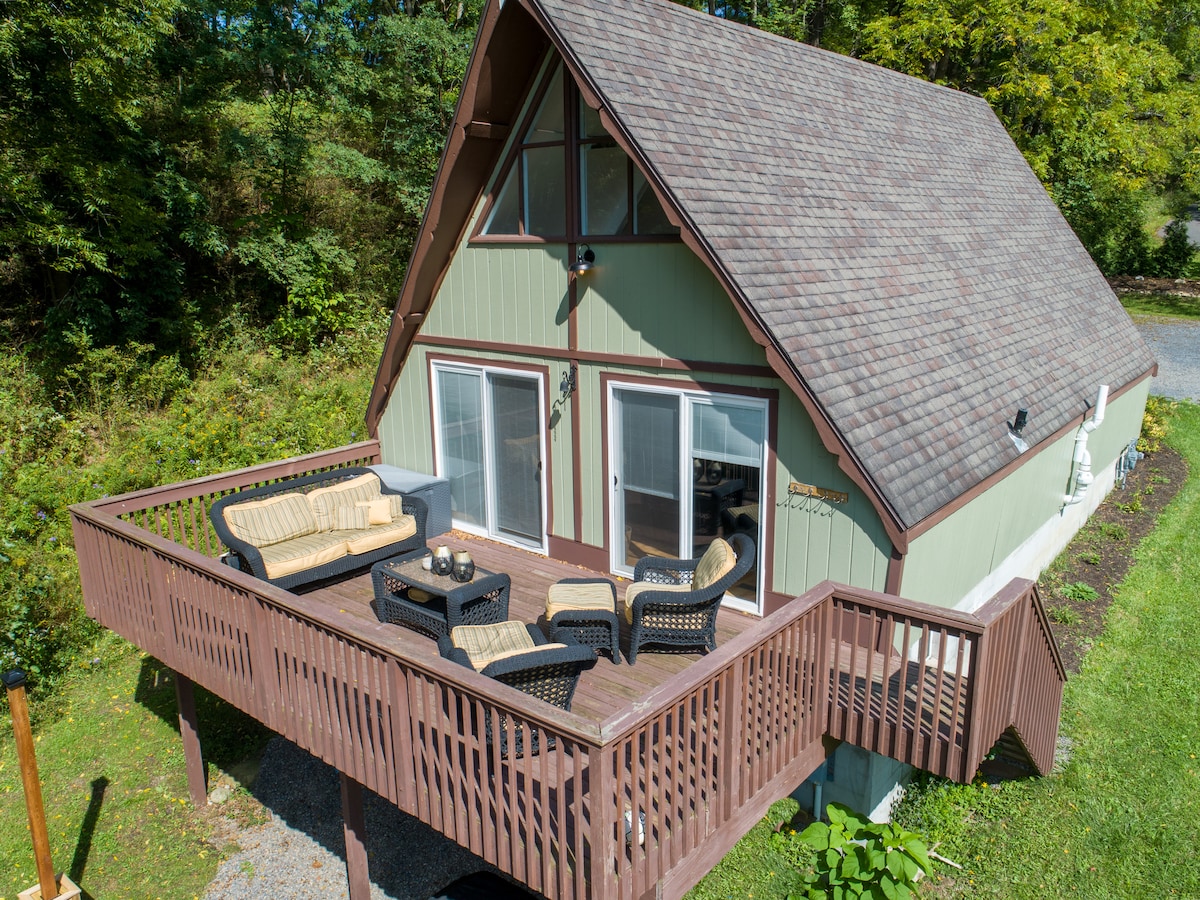 An outdoor deck area is visible, featuring comfortable seating arranged around a small table. The A-frame house exhibits a welcoming façade, complemented by a sloped roof and large glass doors leading indoors. Lush greenery surrounds the property, enhancing the serene atmosphere.