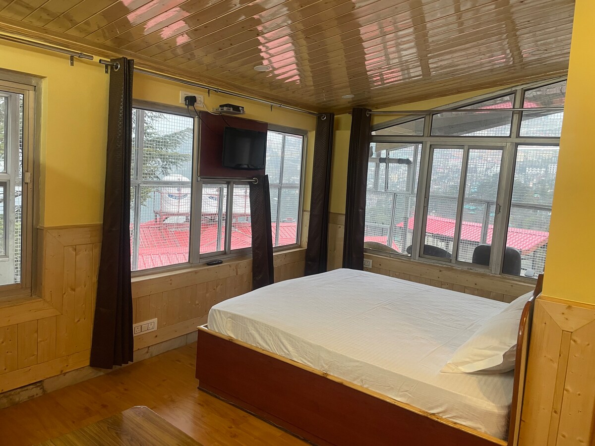 A well-lit sleeping area features a spacious bed with crisp white linens. Large windows offer views of the surrounding area, while curtains frame the windows for privacy. A small wall-mounted television is visible, contributing to a cozy environment.