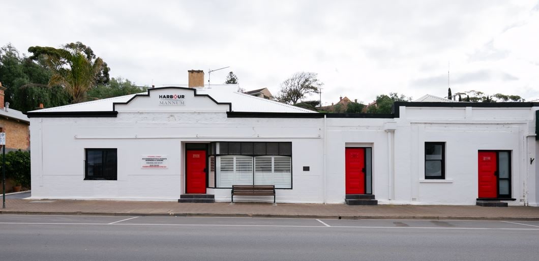 A charming facade of a historic stone building is displayed, featuring a white exterior with red doors. A simple wooden bench is positioned in front, inviting passersby to sit and enjoy the surroundings. The architecture reflects the building's rich history.
