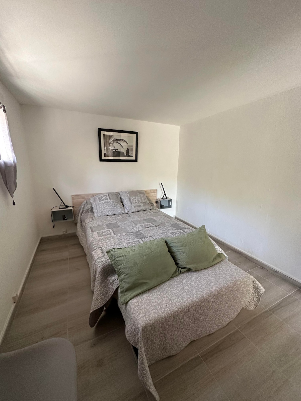 A cozy sleeping area features a spacious bed adorned with a light-colored quilt and green decorative pillows. Two bedside tables with lamps flank the bed, and a piece of wall art adds a subtle touch to the clean, minimalist space.