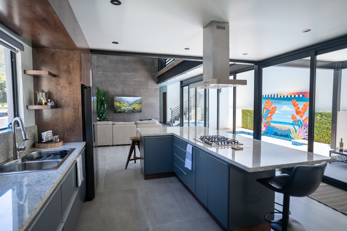An open-plan kitchen features sleek cabinetry and a large granite island with seating. Floor-to-ceiling glass doors provide views of the pool and outdoor area, while a modern living space with a light couch is visible in the background. Artwork decorates a wall, adding color.