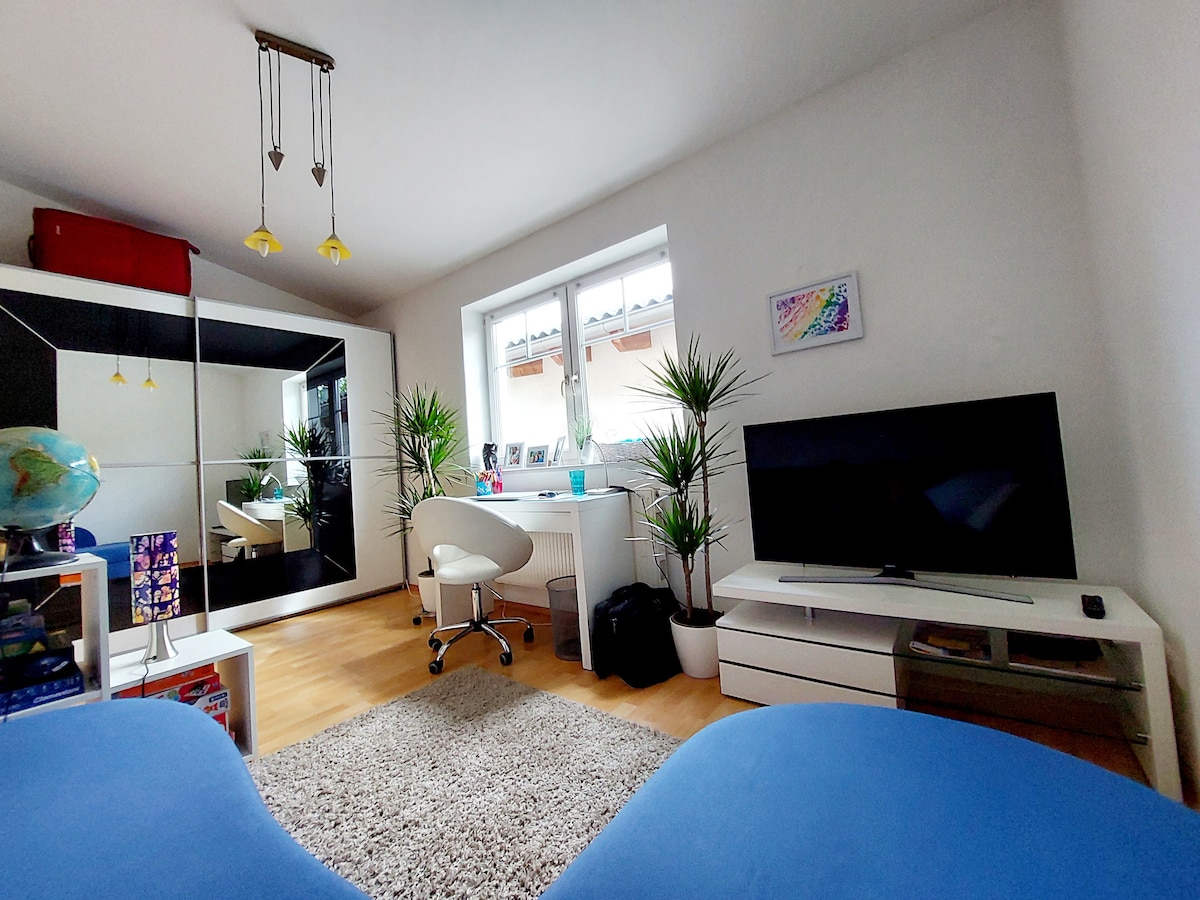 A bright room is visible, featuring a large window allowing natural light to flood in. A workspace with a desk chair and multiple potted plants adds a touch of greenery. A television is mounted on a white media unit, while a soft area rug sits on the wooden floor.
