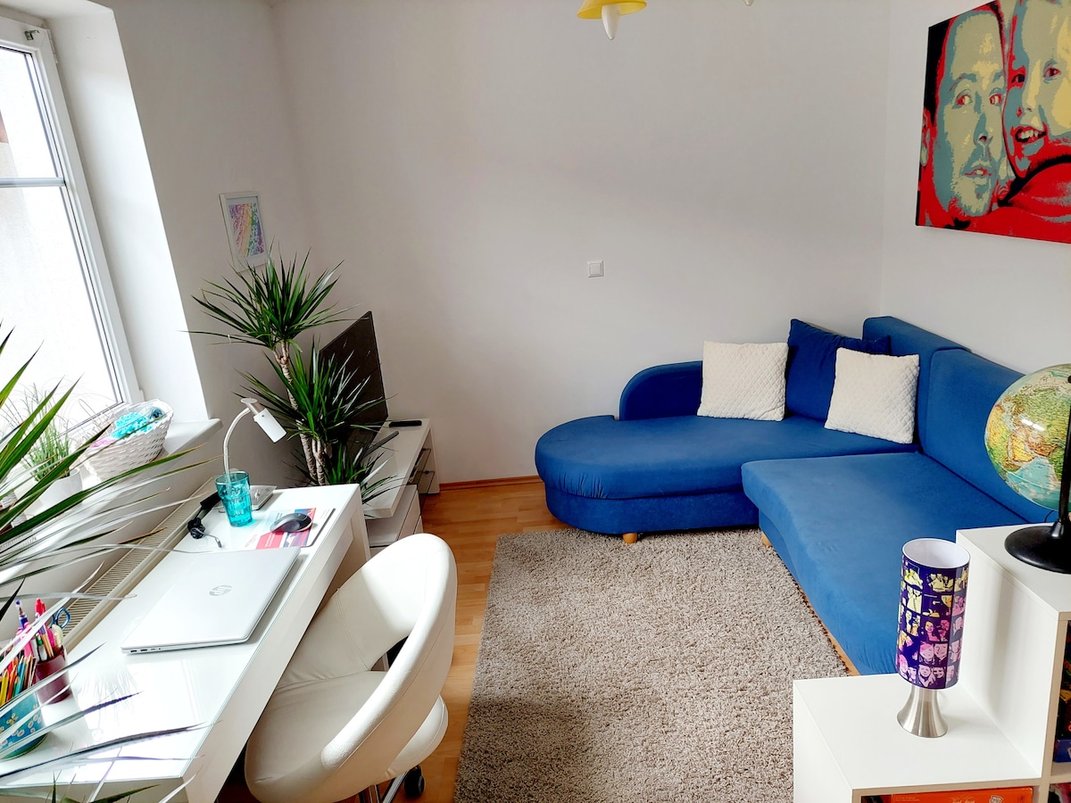 A cozy living area features a blue sectional sofa paired with two white cushions. A desk is positioned against the wall, equipped with a laptop and colorful stationery. A television is visible, and plants add a touch of greenery. Soft light filters through the window.