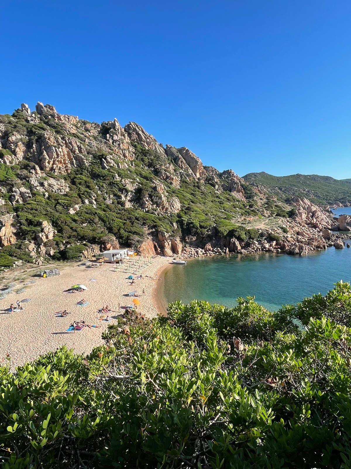 A serene beach scene is depicted, with golden sand gently meeting turquoise waters. Lush green foliage frames the coastline, which is bordered by rugged cliffs. A few beachgoers are visible on the shore, enjoying the tranquil environment under clear blue skies.