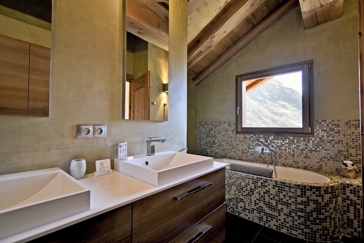 A spacious bathroom features dual sinks with modern fixtures, complemented by a large mirror above each sink. A freestanding bathtub is situated near a window that offers a mountain view. Natural light highlights the earthy tones of the walls and the unique tile design.