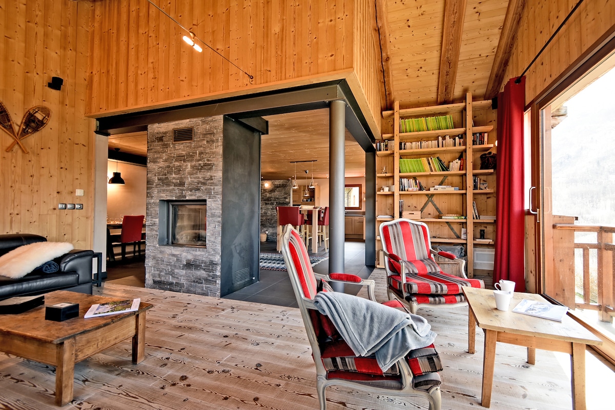 The spacious living area features wooden paneling and a cozy fireplace, creating a warm ambiance. Comfortable seating is arranged with striped chairs and a wooden coffee table. A well-stocked bookshelf and open space leading to the kitchen are visible, complemented by large windows allowing natural light.