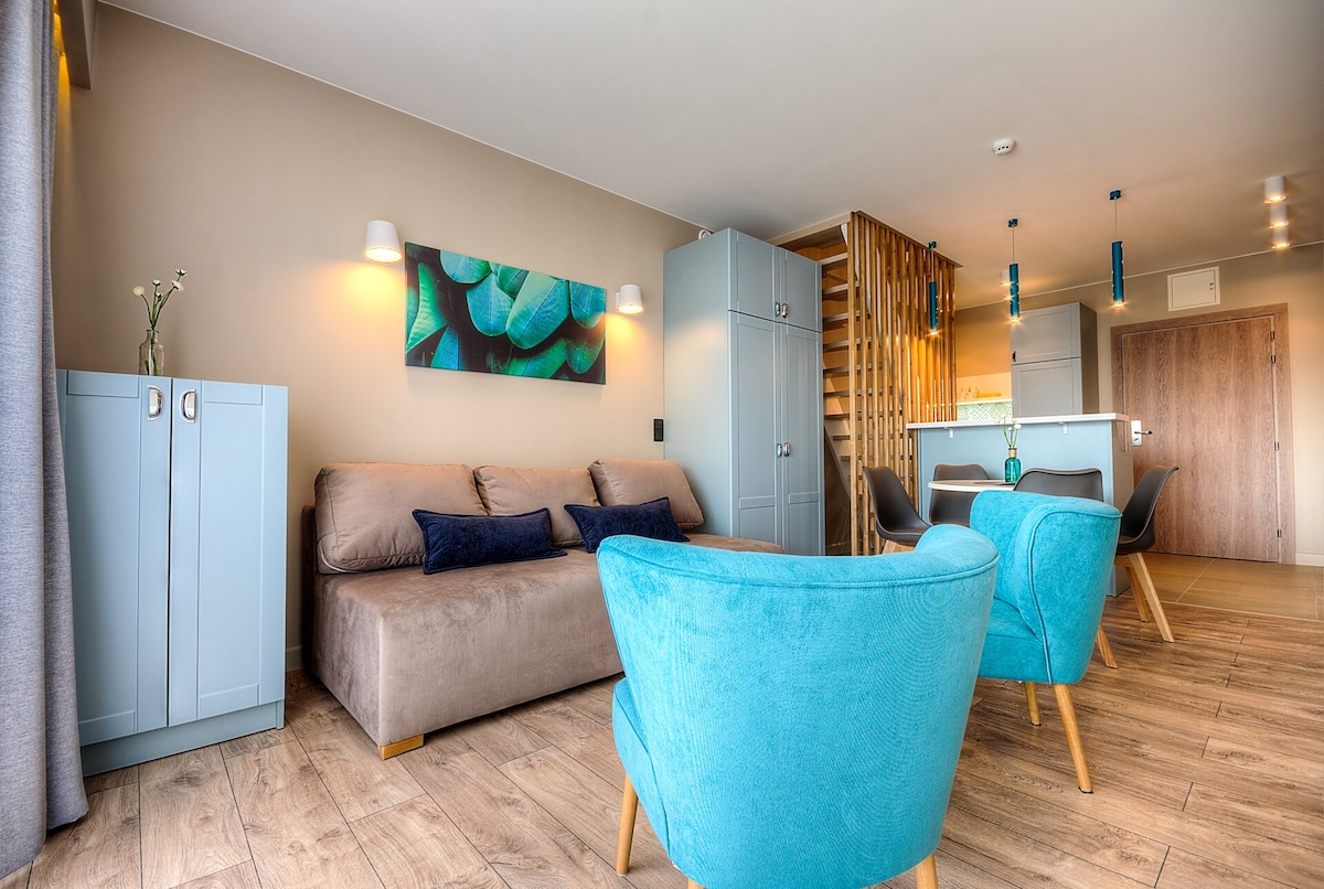 A modern living area is displayed, featuring a comfortable brown sofa and two light blue armchairs. A vibrant piece of wall art is positioned above a light blue cabinet. Brightness is enhanced by natural light filtering through the windows, creating a soothing ambiance.