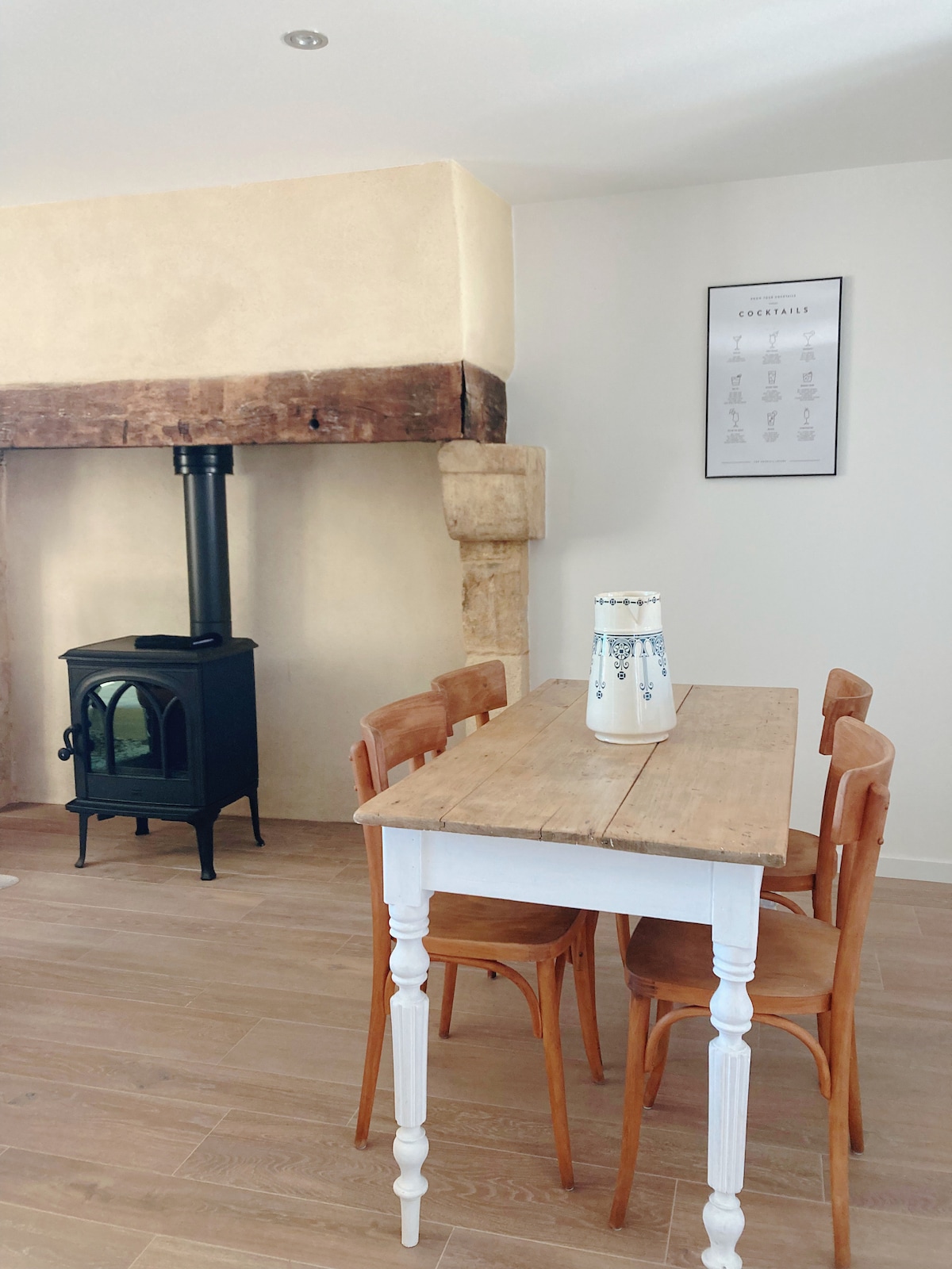 A dining area is presented with a rustic wooden table surrounded by four wooden chairs. A black wood stove adds warmth to the space, complemented by a stone feature wall. A simple decorative vase sits at the center of the table.