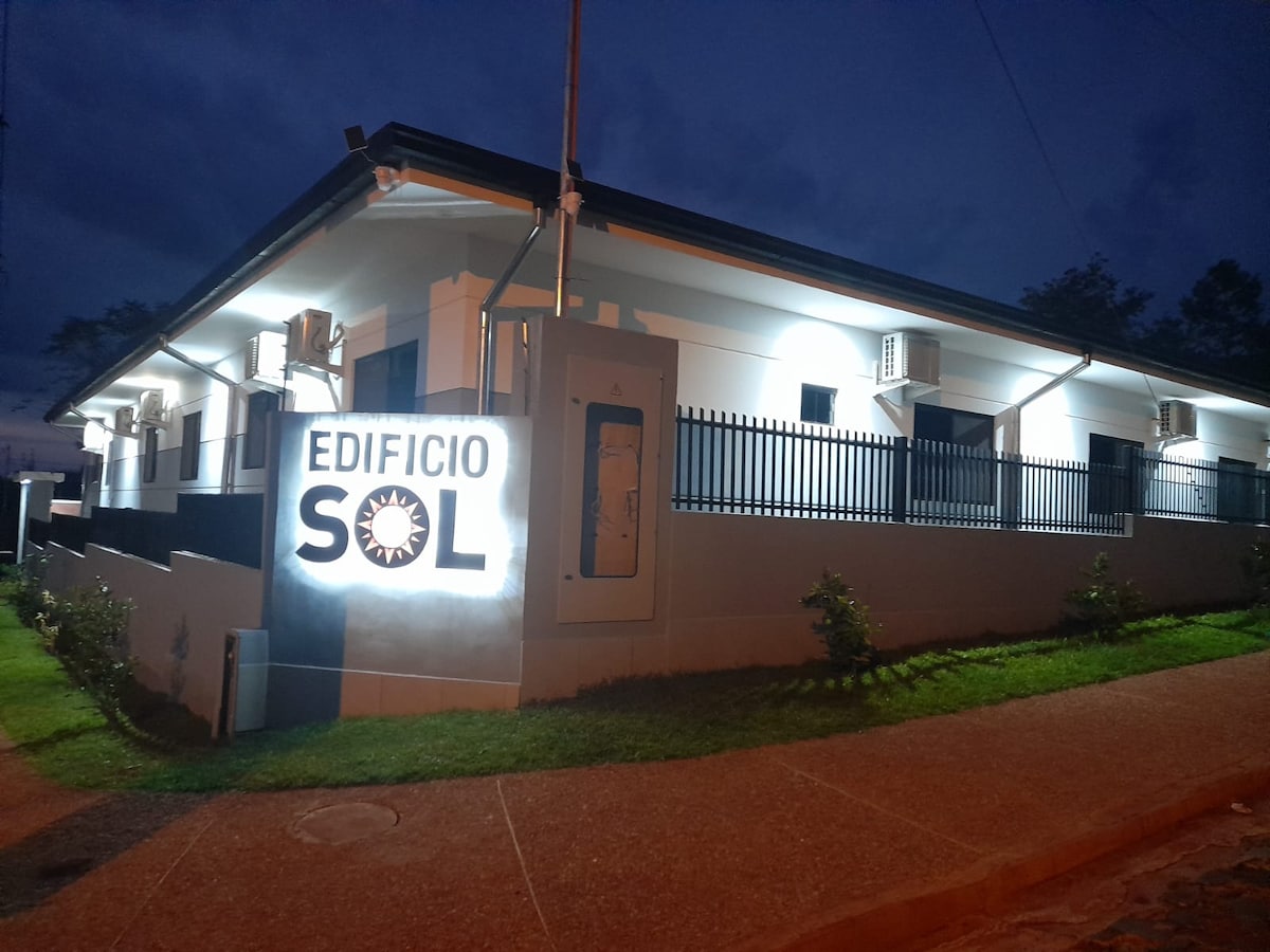 The exterior of the building displays a modern façade, illuminated by exterior lighting. A clear sign reading 'Edificio Sol' is prominently featured. The surrounding landscape includes well-maintained grass and small shrubs, with several air conditioning units visible on the structure.