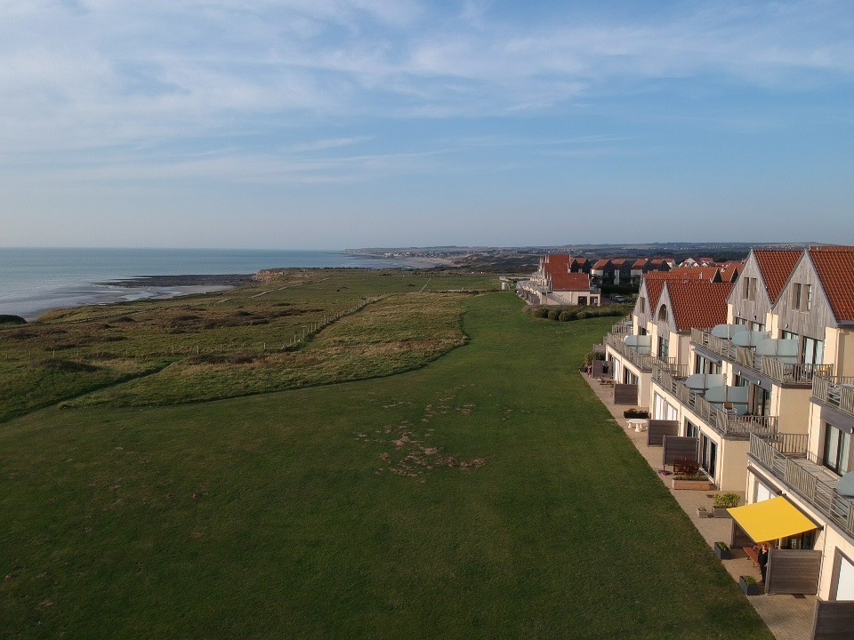 A scenic view captures a stretch of coastline lined with green grass and gentle slopes leading to the sea. The image showcases a series of residential buildings with red-roofed gables, set against a clear blue sky.