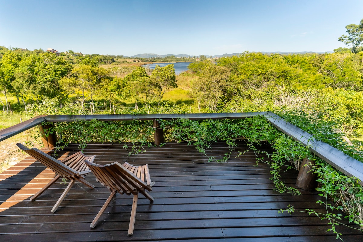 A wooden deck is set with two lounge chairs, surrounded by lush greenery. The tranquil view of the lake and rolling hills is visible in the background, creating a serene space for relaxation.