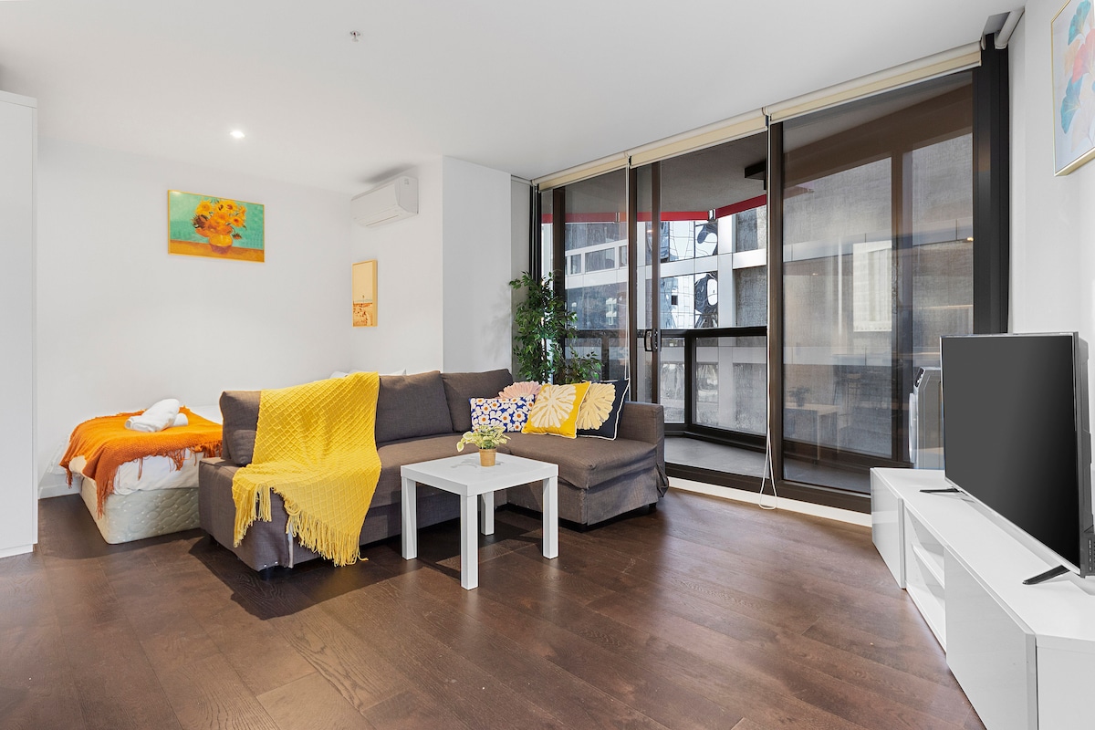 A modern living area features a spacious double-size sofa bed adorned with bright cushions and a cozy throw blanket. A coffee table sits in front of the sofa, with a smart TV positioned on a media unit. Large windows allow natural light to illuminate the room.