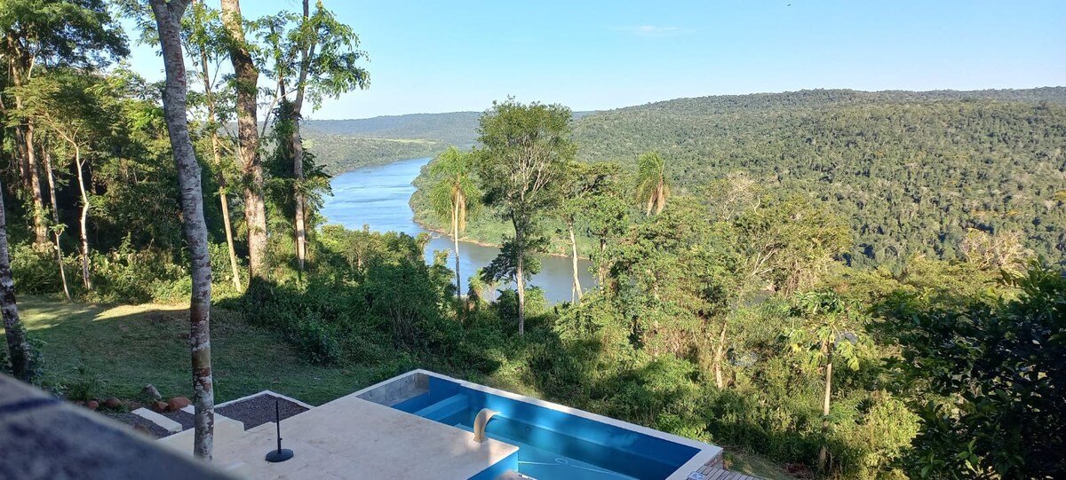 An outdoor area displays a modern pool overlooking a lush landscape. Trees frame the scene, while the river meanders through the greenery in the distance, showcasing the natural beauty of the surrounding jungle.