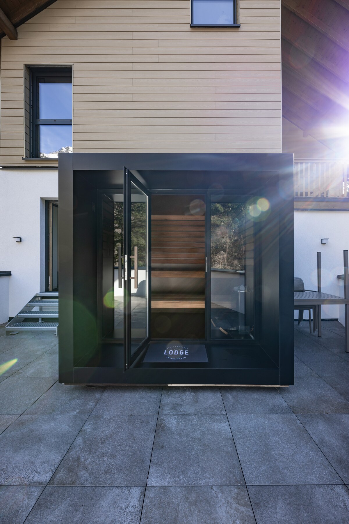 A modern outdoor sauna is positioned on a stone patio, featuring glass walls and a sleek black frame. Sunlight reflects off the surfaces, while the entrance invites access. The structure is complemented by a few chairs nearby, enhancing relaxation in a natural setting.