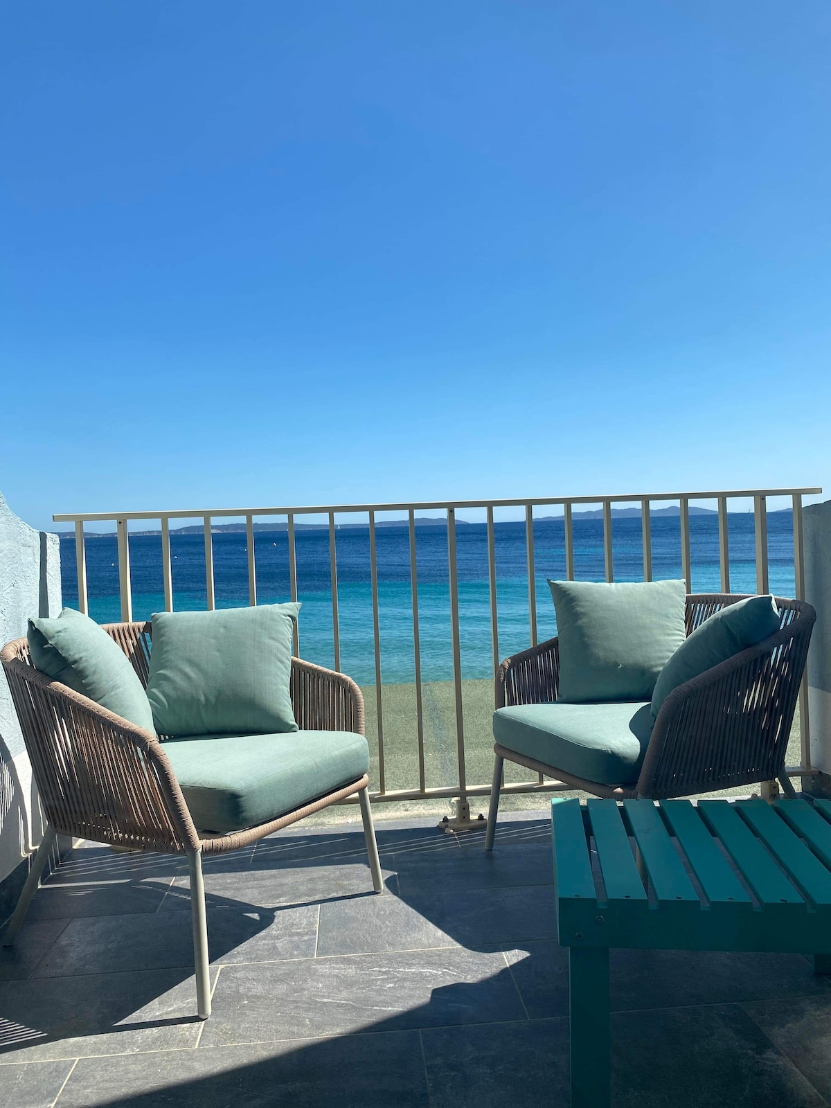 A small balcony features two wicker chairs with soft cushions, arranged against a backdrop of the ocean. The clear blue sky meets the water’s horizon, creating a serene view. A compact table is positioned between the chairs, enhancing the inviting outdoor space.