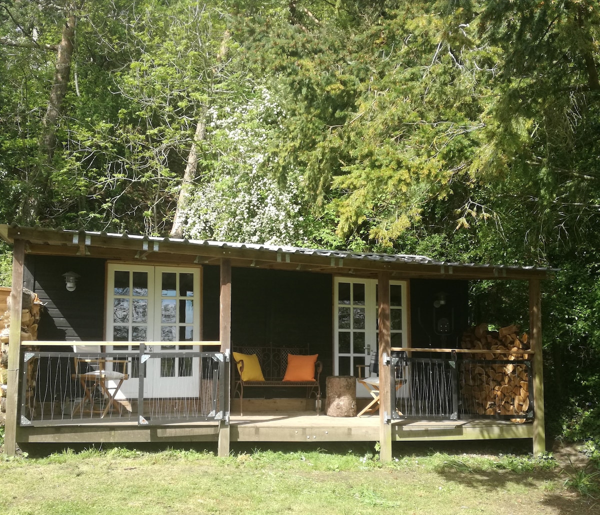 A wooden cabin features a cozy veranda with wooden railings and a stack of firewood to the side. Two sets of double doors open to the living space, which is shaded by lush greenery from surrounding trees. A welcoming orange cushion adds a touch of color.