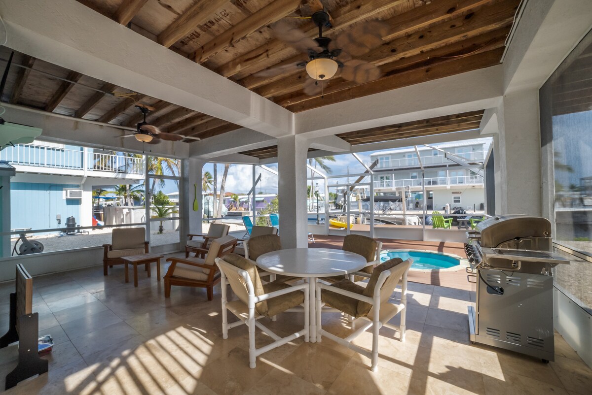 A spacious outdoor patio features a round dining table surrounded by cushioned chairs. A seating area with wooden chairs offers additional relaxation space. A grill is positioned nearby, and a glimpse of the water can be seen through the screened enclosure.