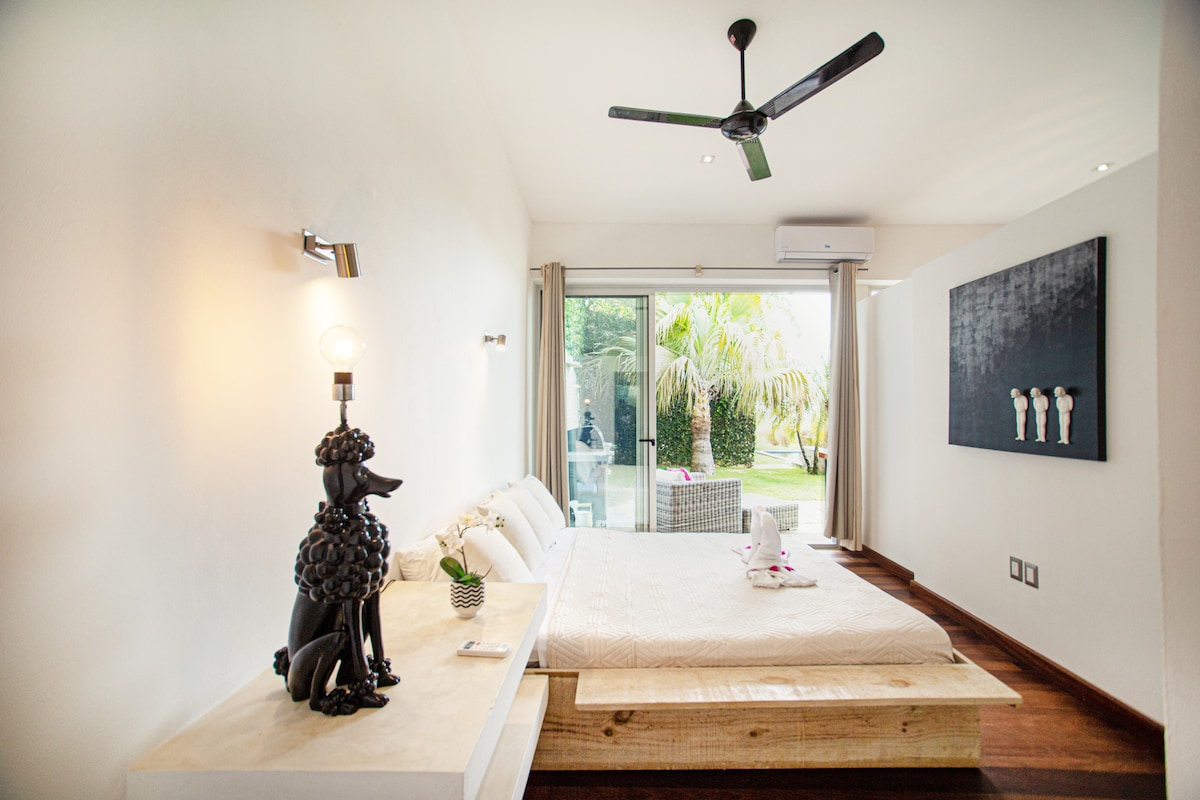 A bright bedroom features a spacious bed with a light wooden frame and a plush headboard. Natural light pours in through sliding glass doors, leading to lush greenery outside. A decorative lamp shaped like a poodle adds a touch of character, while a ceiling fan enhances ventilation.
