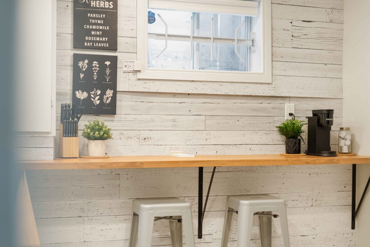 A kitchen area features a light wooden breakfast bar accompanied by two metal stools. Wall-mounted herb illustrations enhance the atmosphere. A coffee maker and a small plant are positioned on the counter, with a window allowing natural light into the space.