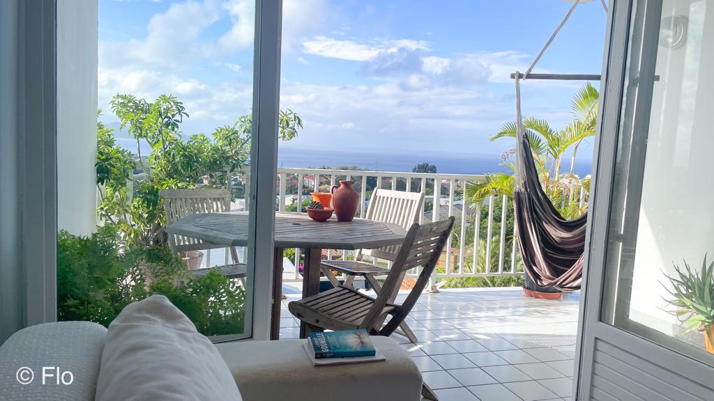 A sunlit terrace features a wooden table with seating for four, overlooking a scenic view of the ocean and surrounding greenery. A hammock sways gently nearby, surrounded by tropical plants, while a soft throw lies on the armrest of a white sofa in the foreground.