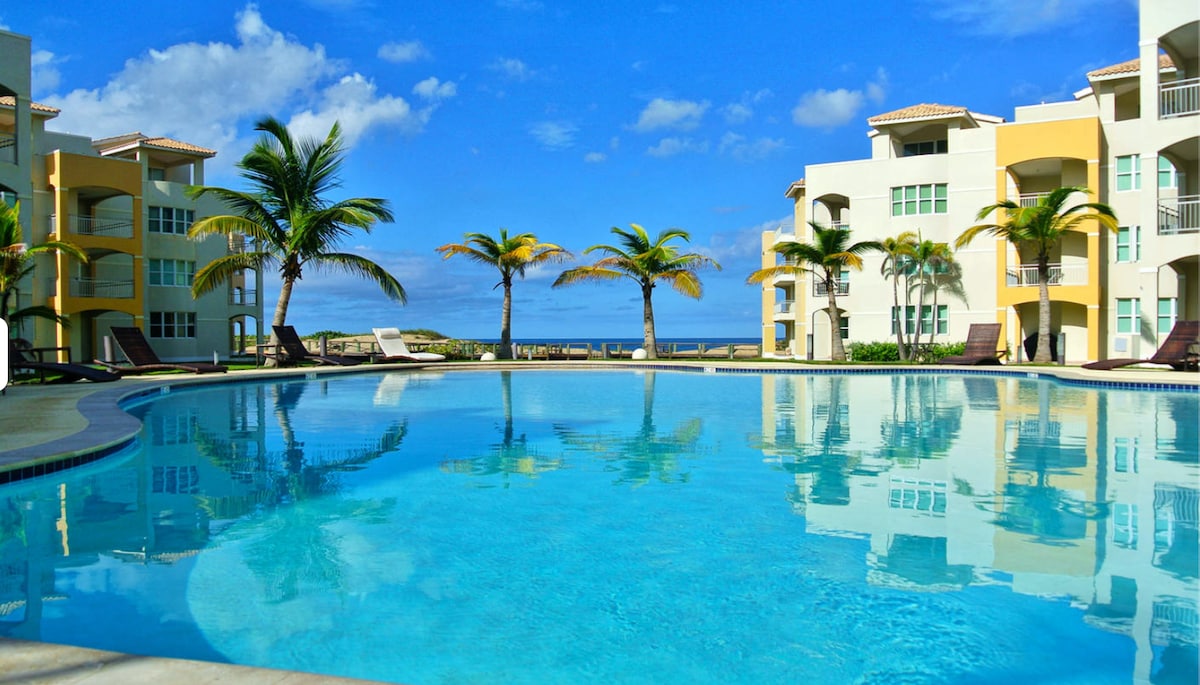 A large swimming pool is framed by palm trees, reflecting the clear blue sky. Surrounding the pool, the beige and yellow buildings create a serene environment, and views of the beach are visible in the distance.