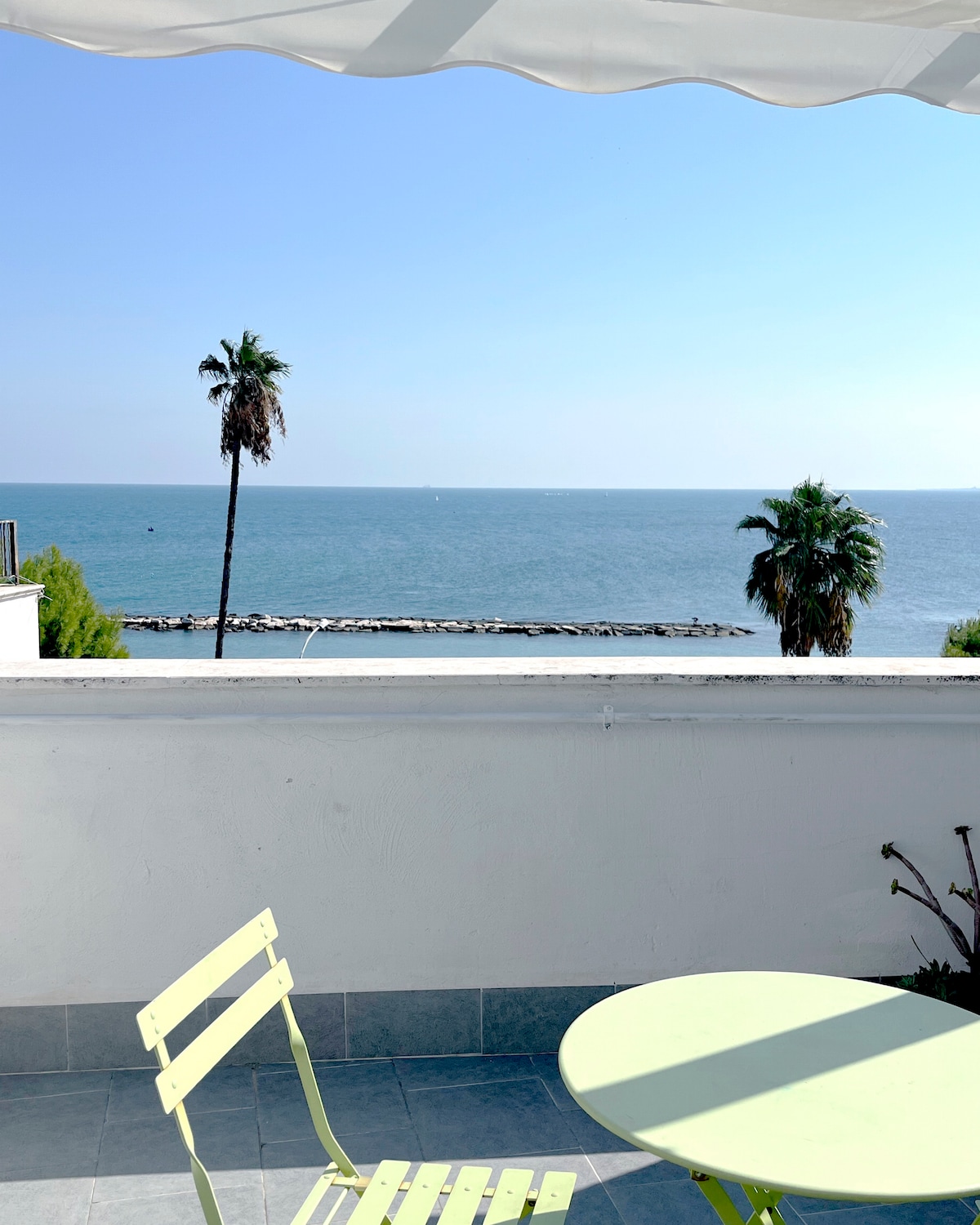 A private terrace offers unobstructed views of the sea, featuring clear blue waters and a distant shoreline. Two green chairs and a round table provide a space for relaxation. Palm trees frame the scene, swaying gently in the breeze under a bright, sunny sky.