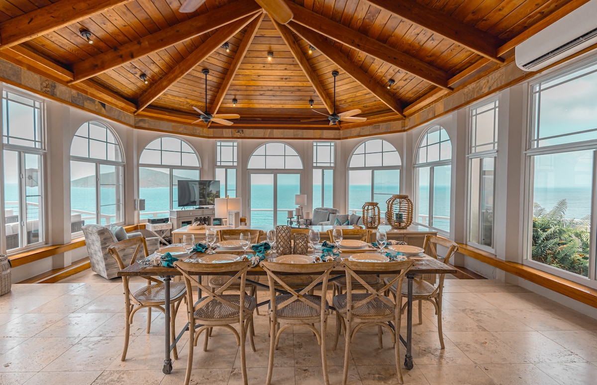 A spacious dining area features a large wooden table surrounded by rattan chairs, set for a meal. Large windows allow natural light and showcase ocean views, while a high, wooden ceiling with fans creates an airy atmosphere.