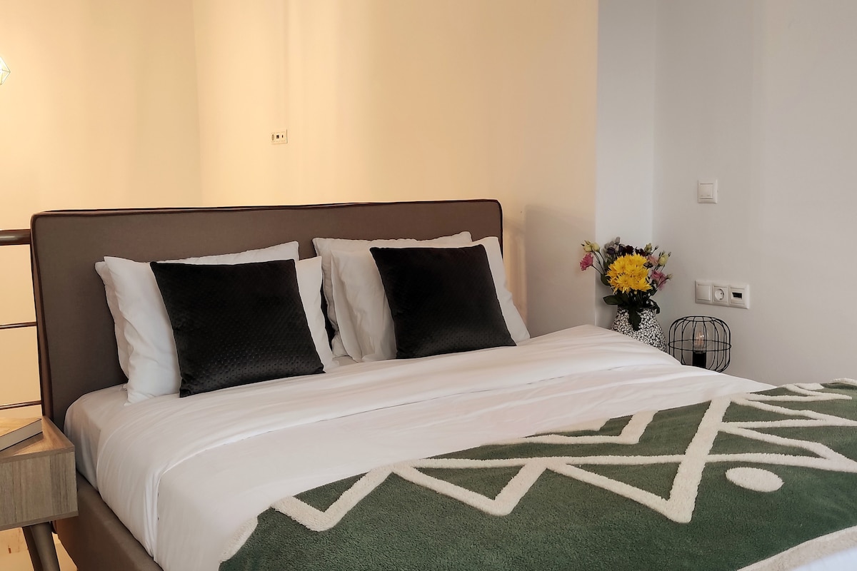 A spacious bed is dressed with white linens and accented by dark pillows. A decorative green blanket with geometric patterns is laid at the foot of the bed. A bouquet of flowers sits in a vase nearby, adding a touch of color to the neutral wall.