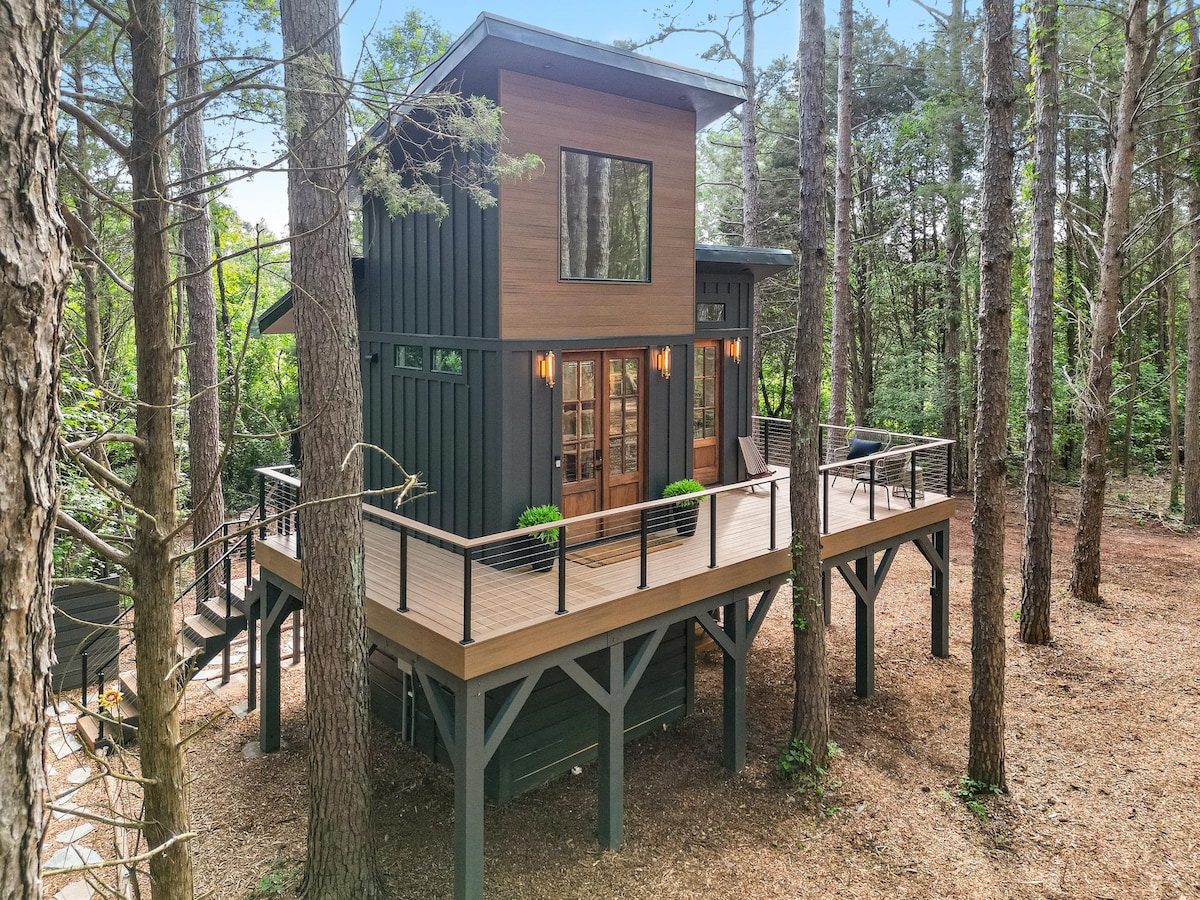 A modern treehouse stands tall among the trees, featuring large windows that invite natural light. A spacious wraparound deck is adorned with stylish railings and greenery, enhancing the connection to the surrounding forest.