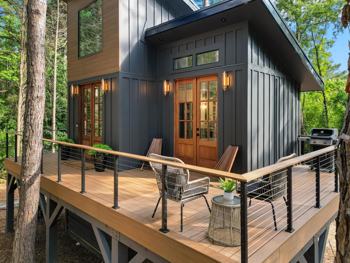 An elegant treehouse features a spacious deck surrounded by trees, showcasing a blend of modern design and natural elements. Two outdoor chairs and a small table invite relaxation, while double doors provide access to the interior. A grill is positioned nearby for outdoor cooking.