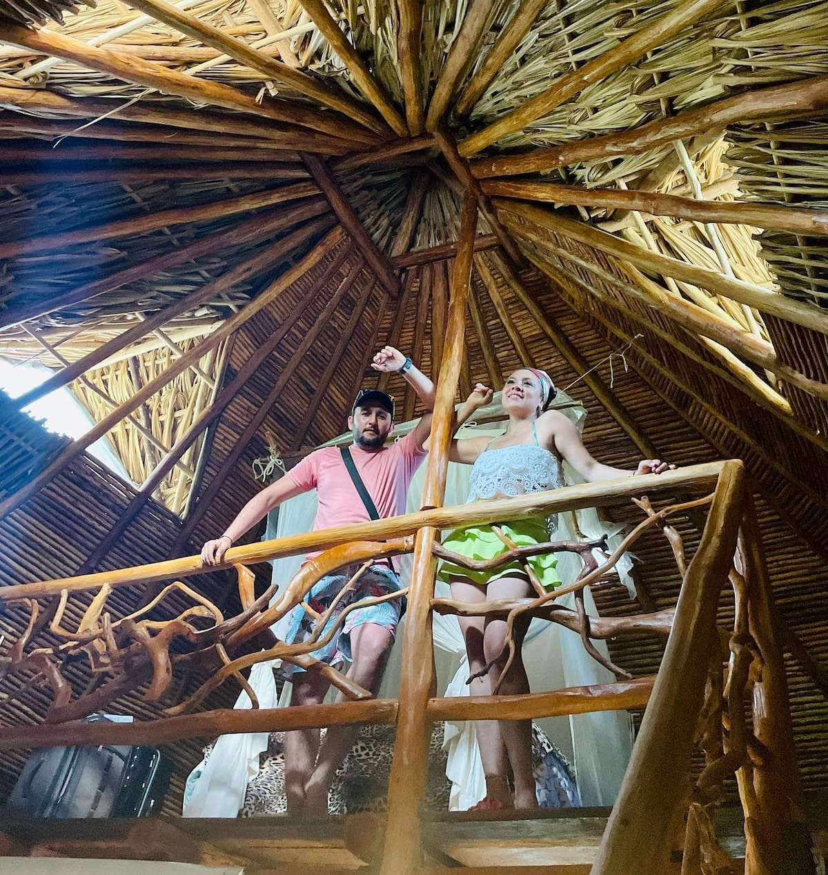 An open palapa-style ceiling showcases intricate wooden beams and thatched roofing. Two individuals stand on a wooden balcony, with natural light streaming through the upper windows, creating a bright and airy ambiance in the treehouse-like space.