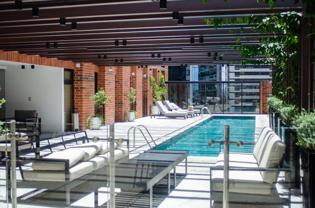 An outdoor pool area is presented with sleek lounge chairs and a shaded seating arrangement. The pool features clear water, surrounded by lush greenery and stylish brick walls, with modern architecture visible in the background.