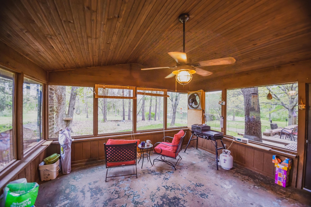 A screened-in porch features a wooden ceiling and large windows overlooking a wooded area. Two cushioned chairs are positioned around a small table, with a standing fan providing airflow. A grill is located in one corner, enhancing outdoor cooking options.