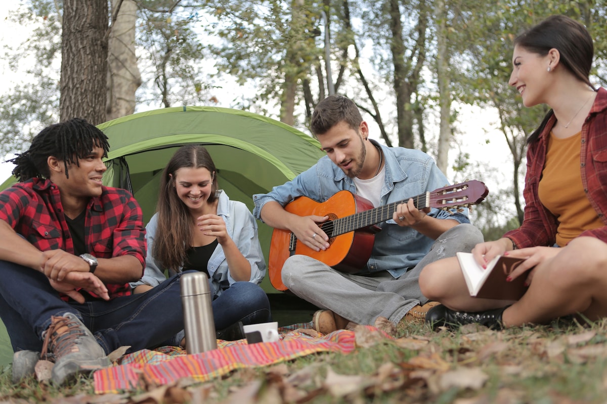 A cozy gathering is depicted outside a green tent surrounded by trees. Four individuals are seen enjoying the outdoors, with one person playing a guitar. A picnic blanket is spread on the ground, and a thermos can be observed nearby.