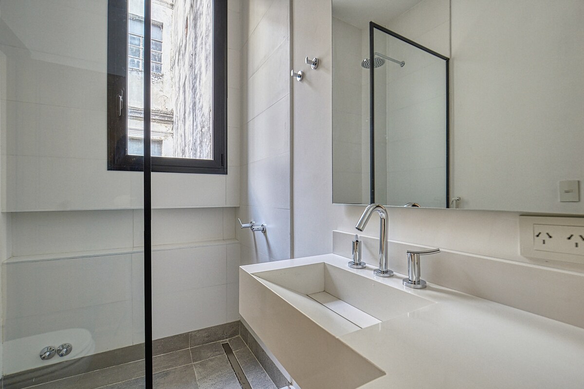 A modern bathroom features a sleek, rectangular sink with three faucets. A large mirror reflects the natural light from a window. Minimalist design elements are present, with white tiled walls and a glass shower enclosure, all contributing to a clean and spacious aesthetic.