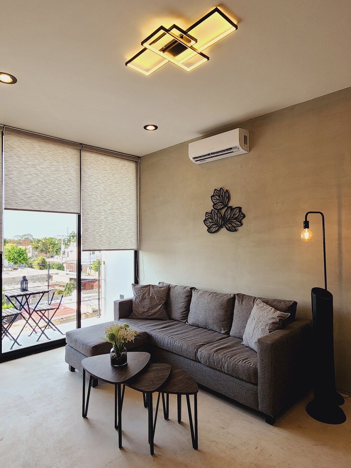 A modern living area is presented with a gray sofa featuring multiple decorative pillows. Two nested tables are placed in front of the sofa. A stylish floor lamp provides soft lighting, and large windows allow natural light to flood the space, complemented by light-filtering shades.