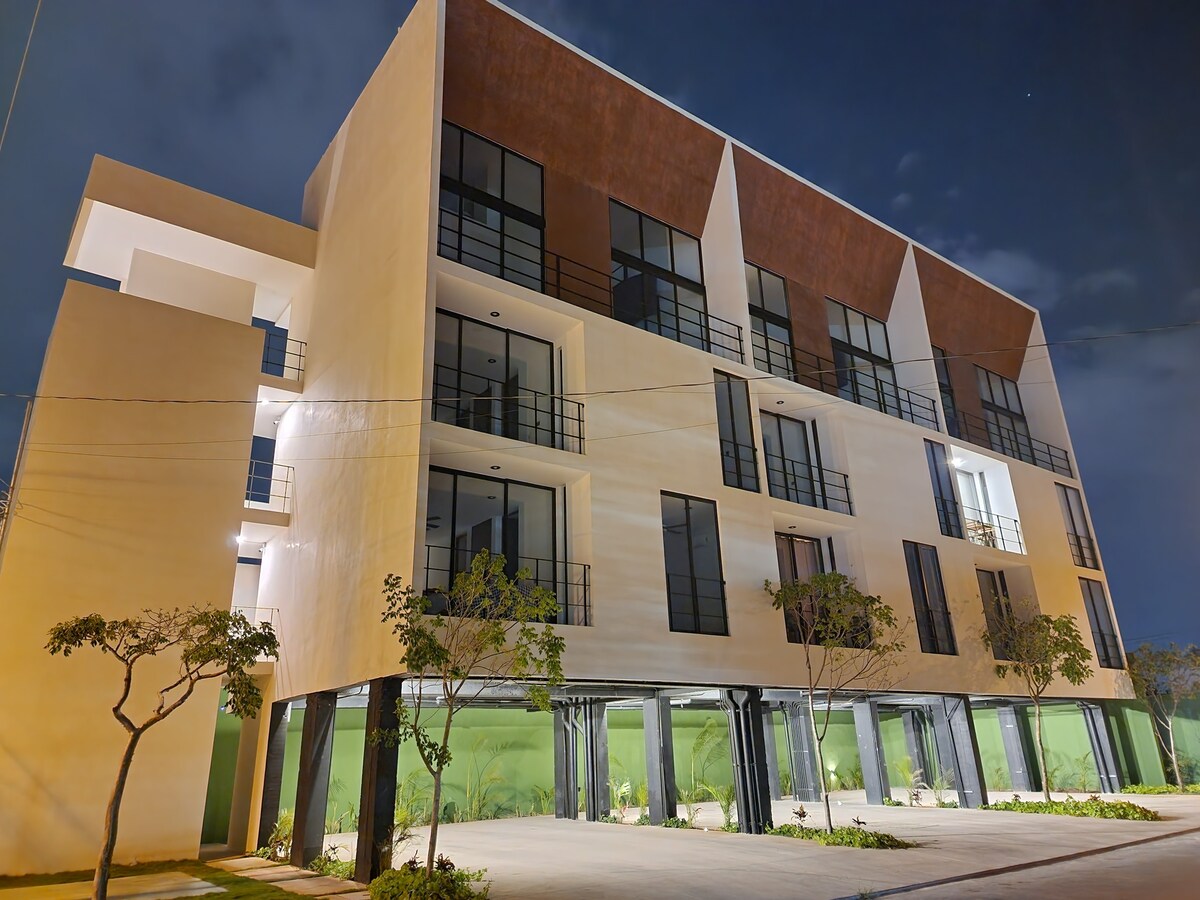 The modern four-story building features a blend of concrete and wood materials, with multiple large glass windows and balconies on each level. The ground is lined with trees and green landscaping, creating a serene environment under the night sky.