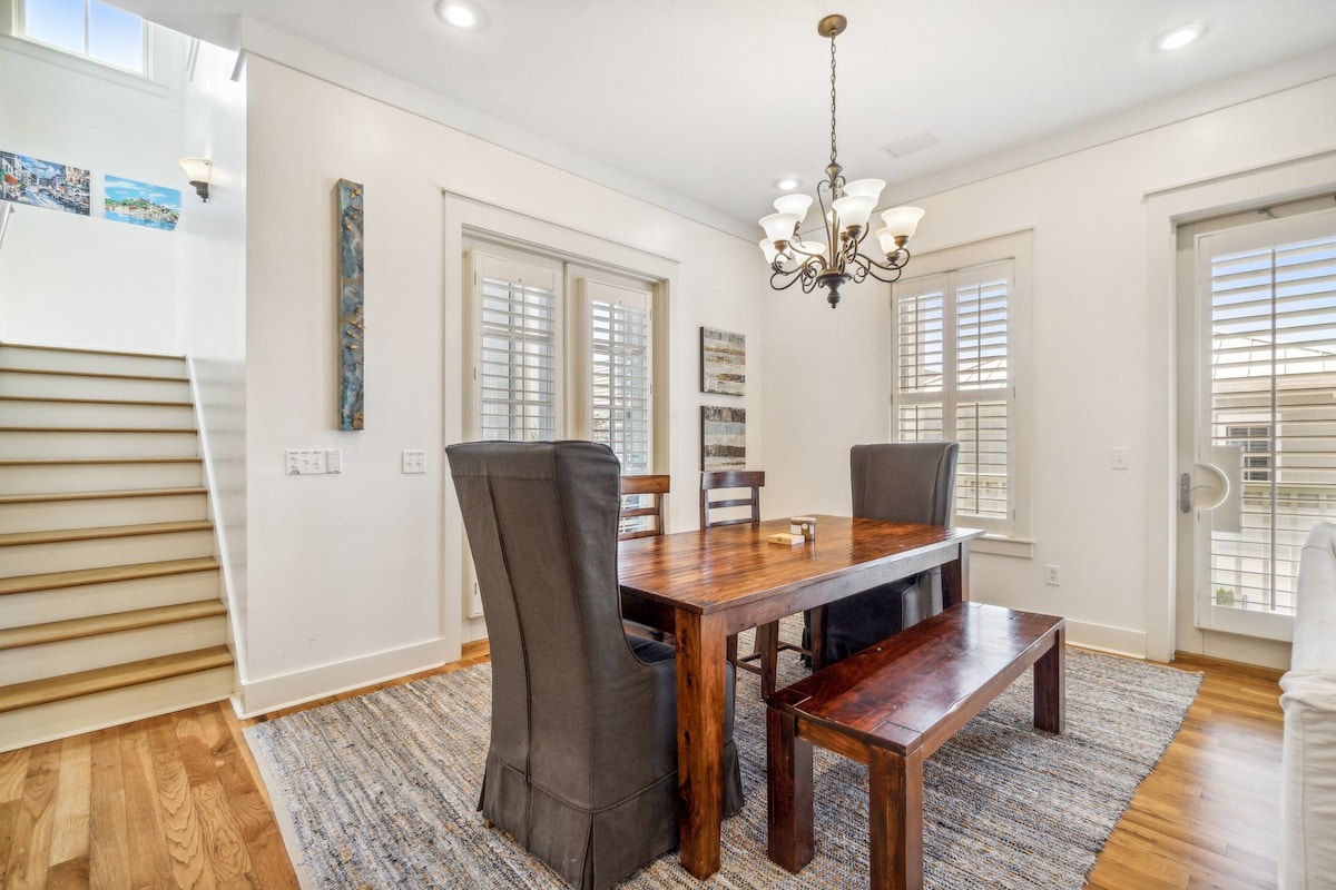 A warm dining area is showcased with a large wooden table surrounded by dark chairs and a matching bench. Light filters through plantation shutters, illuminating the space. A decorative light fixture hangs above, while the wooden staircase is visible in the background.