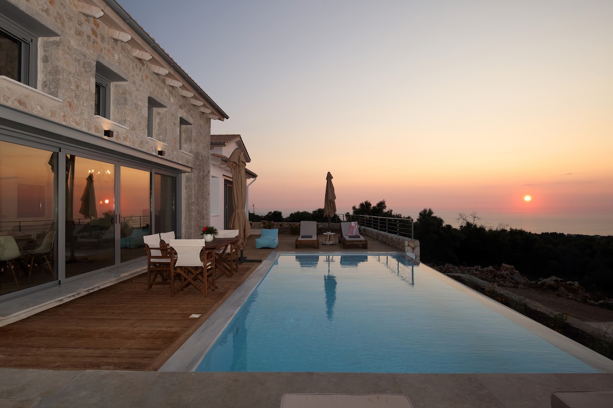 An outdoor space features a private pool reflecting the colors of sunset. The wooden decking accommodates seating areas with stylish chairs and tables. A glimpse of lush greenery surrounds the area, while the villa’s stone exterior is visible under a soft evening light.