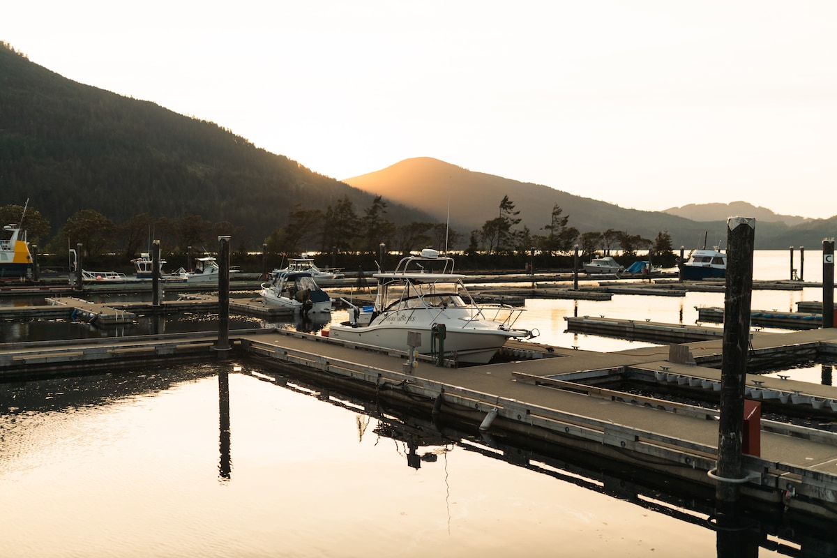 A serene marina at sunset is depicted, with calm waters reflecting the fading light. Several boats are moored along the docks, while gentle hills outline the background. The soft glow of the setting sun casts a warm hue over the scene, enhancing the tranquil atmosphere.