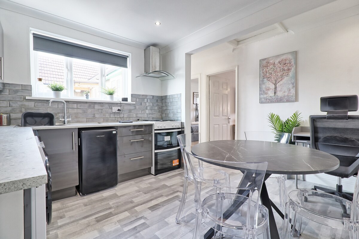 A modern kitchen is equipped with sleek appliances and a stone countertop. A round dining table with transparent chairs complements the space. Natural light is welcomed through a window above the sink, and a potted plant adds a touch of greenery.