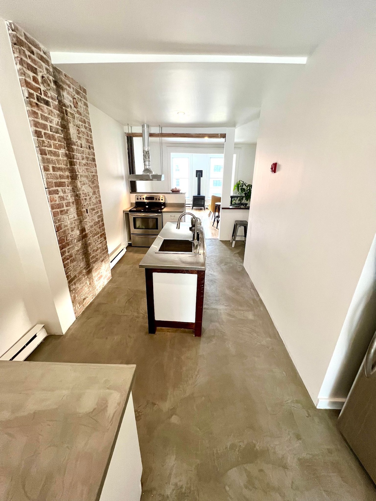 An open kitchen area features a central island with seating, complemented by stainless steel appliances. Exposed brick accents one wall, adding texture to the space. Natural light enters through a nearby window, illuminating the clean lines and modern finishes of the room.