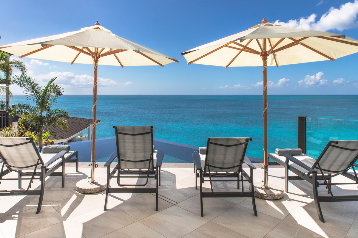 A spacious outdoor terrace features several loungers arranged beneath large umbrellas, overlooking a vibrant turquoise ocean. An infinity pool seamlessly merges with the water, enhancing the serene beachfront views.