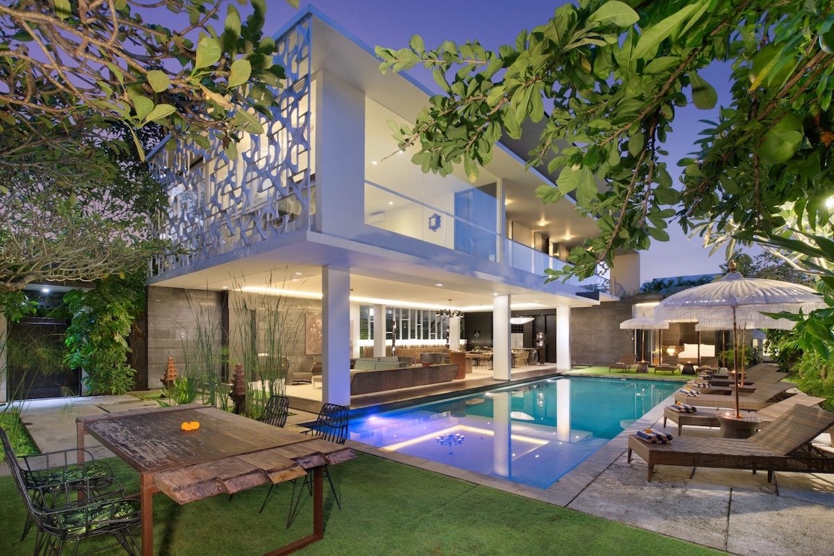 A modern two-story villa is surrounded by lush greenery, featuring a spacious outdoor area and a striking pool reflecting the twilight sky. A wooden dining table with chairs is positioned nearby, while lounge chairs offer a place to relax under umbrellas, completing the tranquil setting.