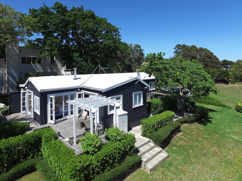 The exterior of a dark-clad house is showcased, surrounded by lush green hedges and trees. A spacious deck with outdoor seating is visible, leading to stone steps that descend into the garden. The clear blue sky emphasizes the inviting outdoor space.