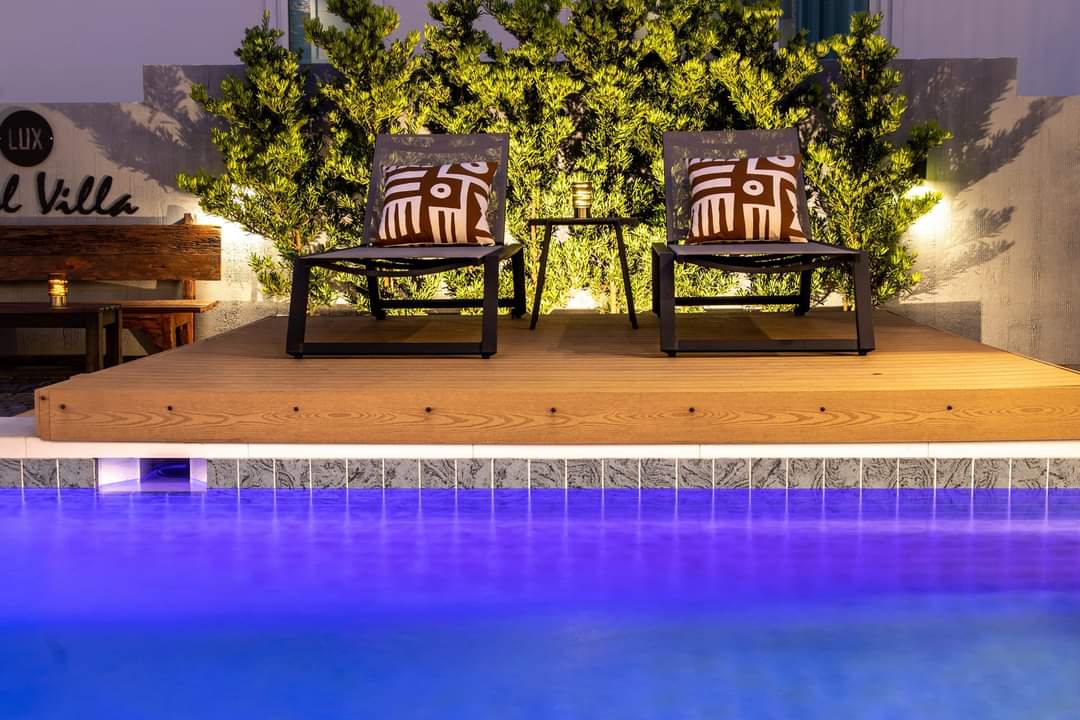 A wooden deck is seen alongside a private swimming pool, featuring two lounge chairs adorned with patterned cushions. Soft lighting casts a gentle glow from the surrounding greenery, creating a calming atmosphere for relaxation.