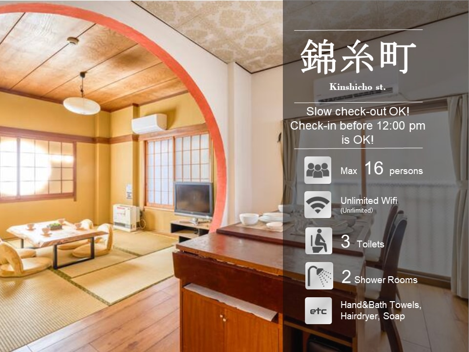 A spacious common area is presented, featuring traditional tatami flooring and a large wooden table surrounded by cushions. Soft natural light enters through large windows, creating a calm environment. The layout accommodates social gatherings, with additional seating options and a view of the kitchenette area.