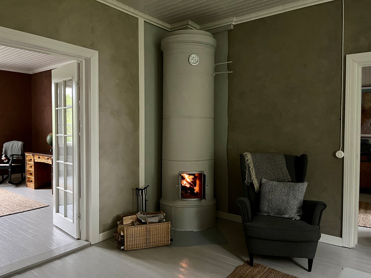 A cozy seating area features a grey potbelly stove, radiating warmth. Nearby, a comfortable armchair is positioned next to the stove, with a patterned cushion inviting relaxation. Natural light filters through the partially open door leading to another room, enhancing the room's welcoming atmosphere.