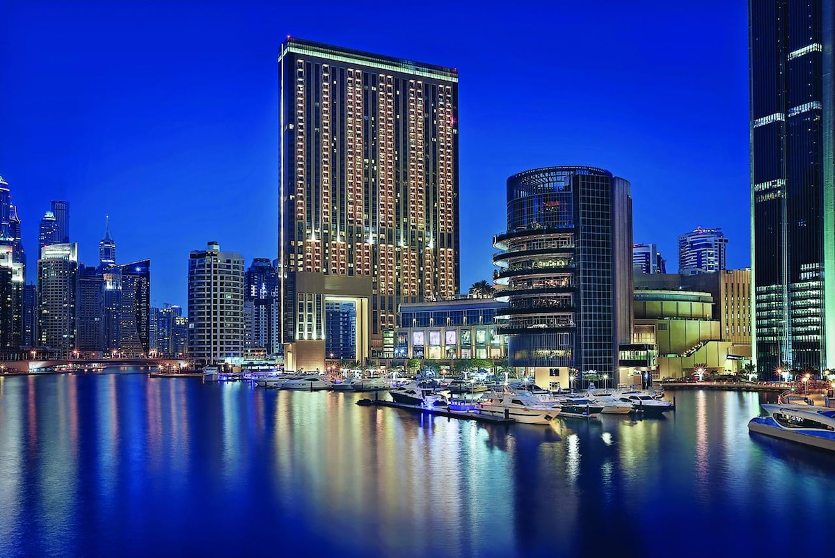 A night view showcases modern architecture along the marina, with illuminated buildings reflecting in the water. Yachts are docked in the foreground, surrounded by a vibrant city skyline, creating a serene ambiance under a deep blue sky.